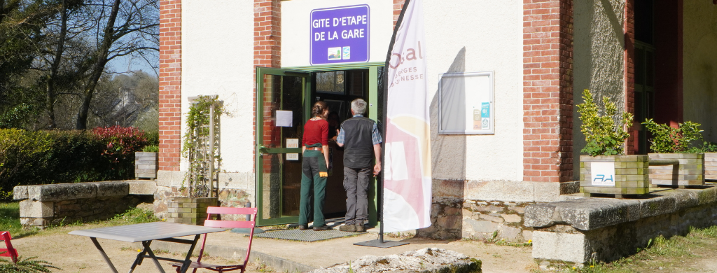Entrée du Gîte d’étape de la Gare dans les Monts d’Arrée avec voyageurs accueillis devant le bâtiment Ostal sous le soleil breton.